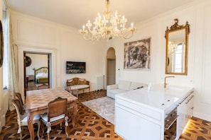 Ein Zimmer mit Esstisch, Stühlen, einer Couch, einem weißen Waschbecken, einer Kristallleuchter, einem Gemälde und einem Spiegel.
