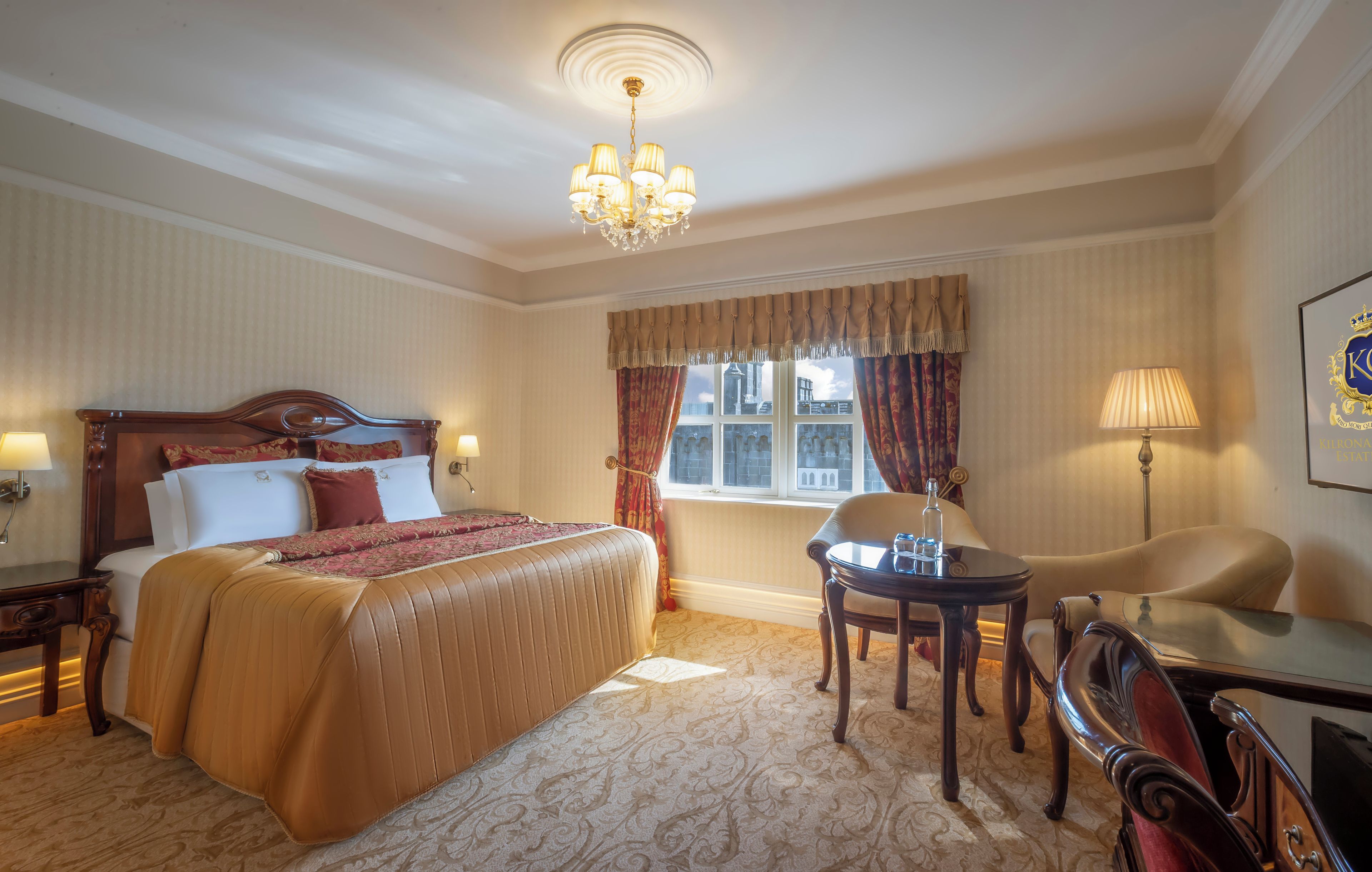A hotel room with a large bed, a chandelier, a window with curtains, a desk, and a chair.