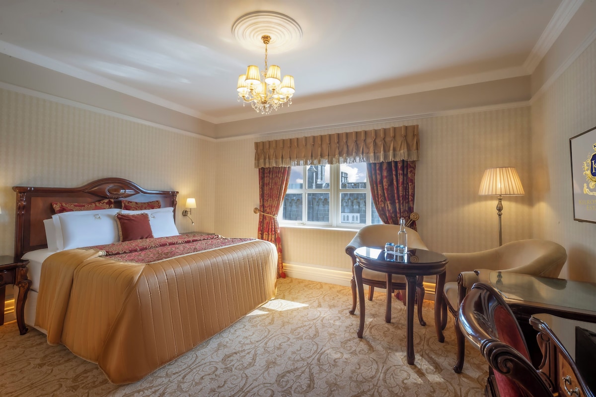 Una habitación de hotel con una cama grande, un candelabro, una ventana con cortinas, un escritorio y una silla.
