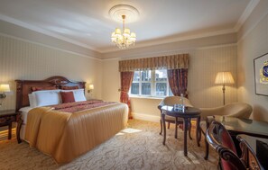 Una habitación de hotel con una cama grande, un candelabro, una ventana con cortinas, un escritorio y una silla.