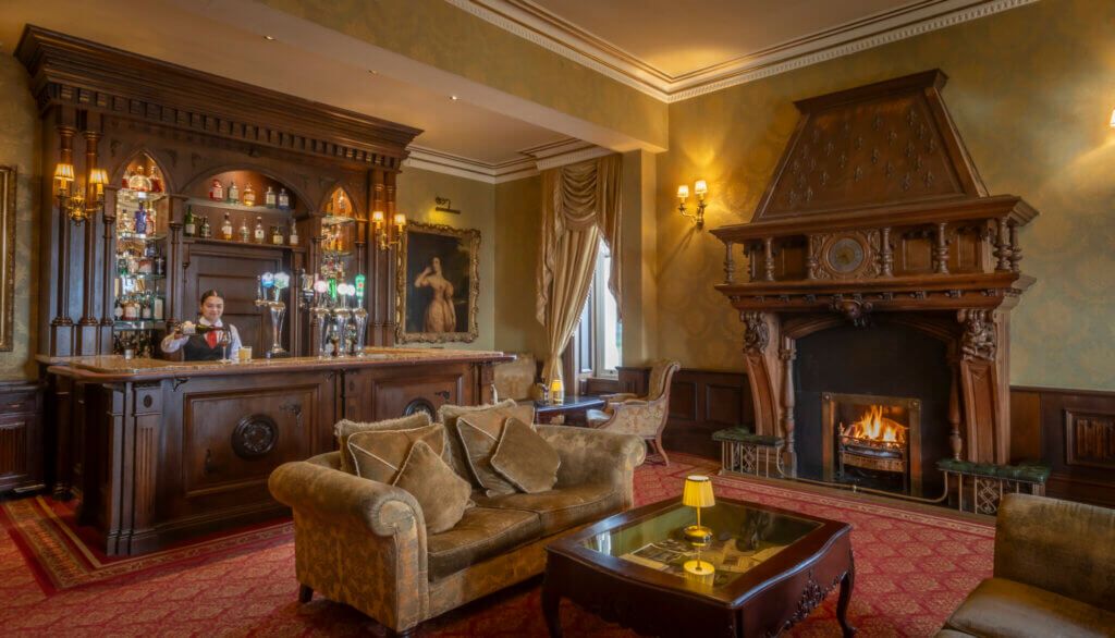 Una zona de bar con un cantinero, una chimenea y un sofá en una sala con detalles de madera labrada y alfombra roja.