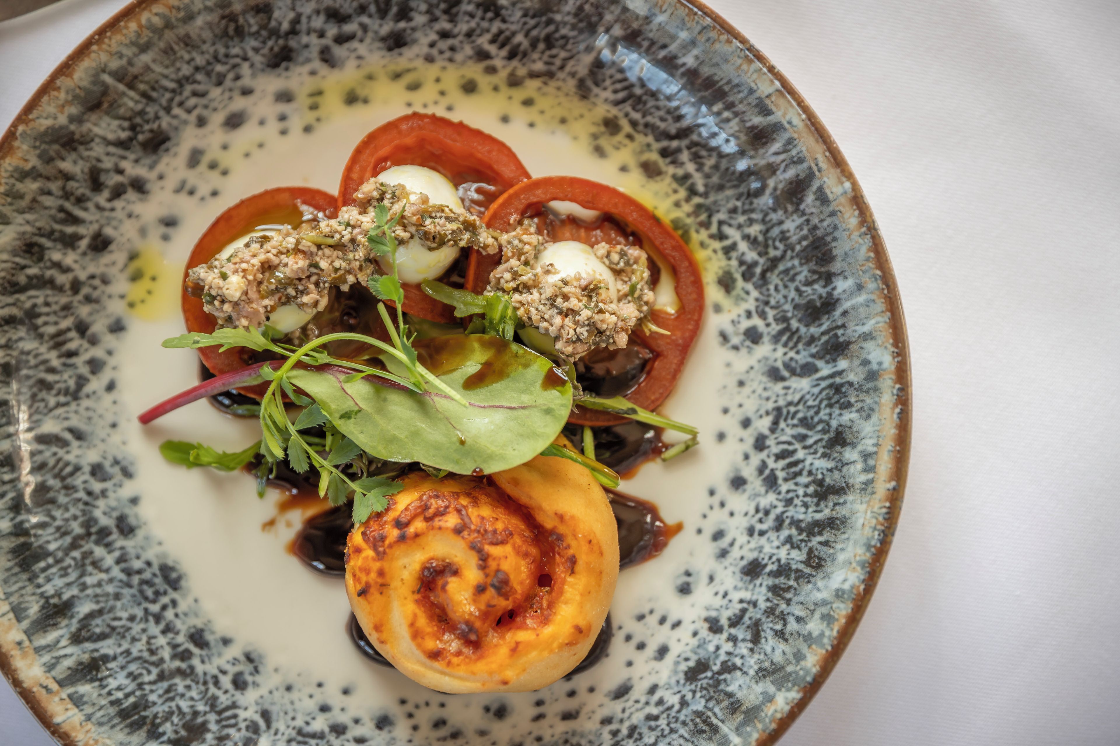 Un plato de comida con diversos ingredientes, incluyendo tomates, verduras de hoja y un elemento redondo que parece ser queso o huevo.
