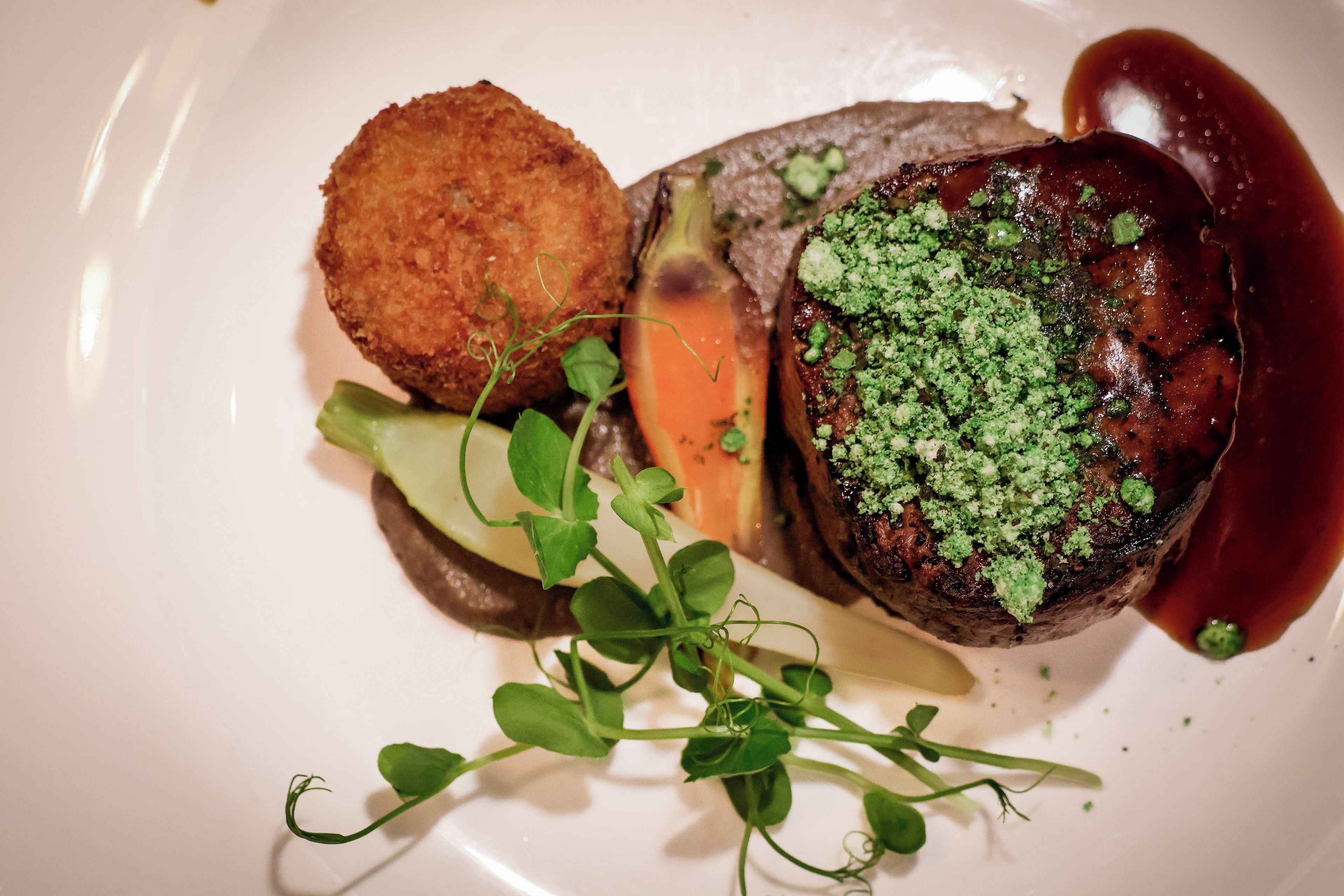 A plated dish with a meat centerpiece, a fried item, and a garnish of green leaves.