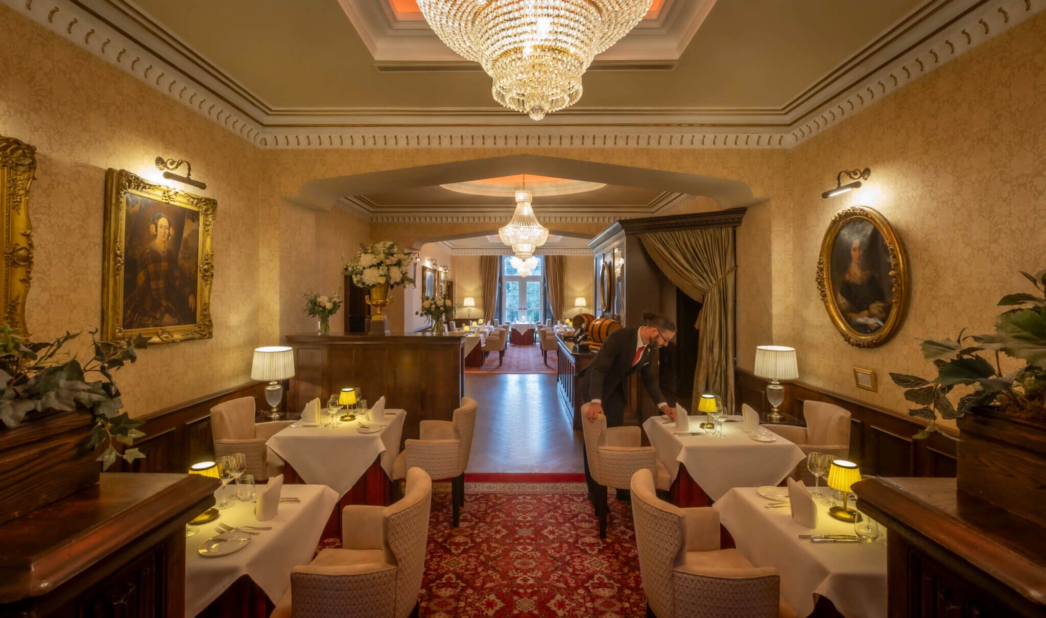 A hotel lobby with a grand chandelier, ornate wall decorations, and a central corridor leading to a dining area.