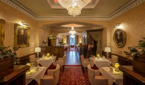 A hotel lobby with a grand chandelier, ornate wall decorations, and a central corridor leading to a dining area.