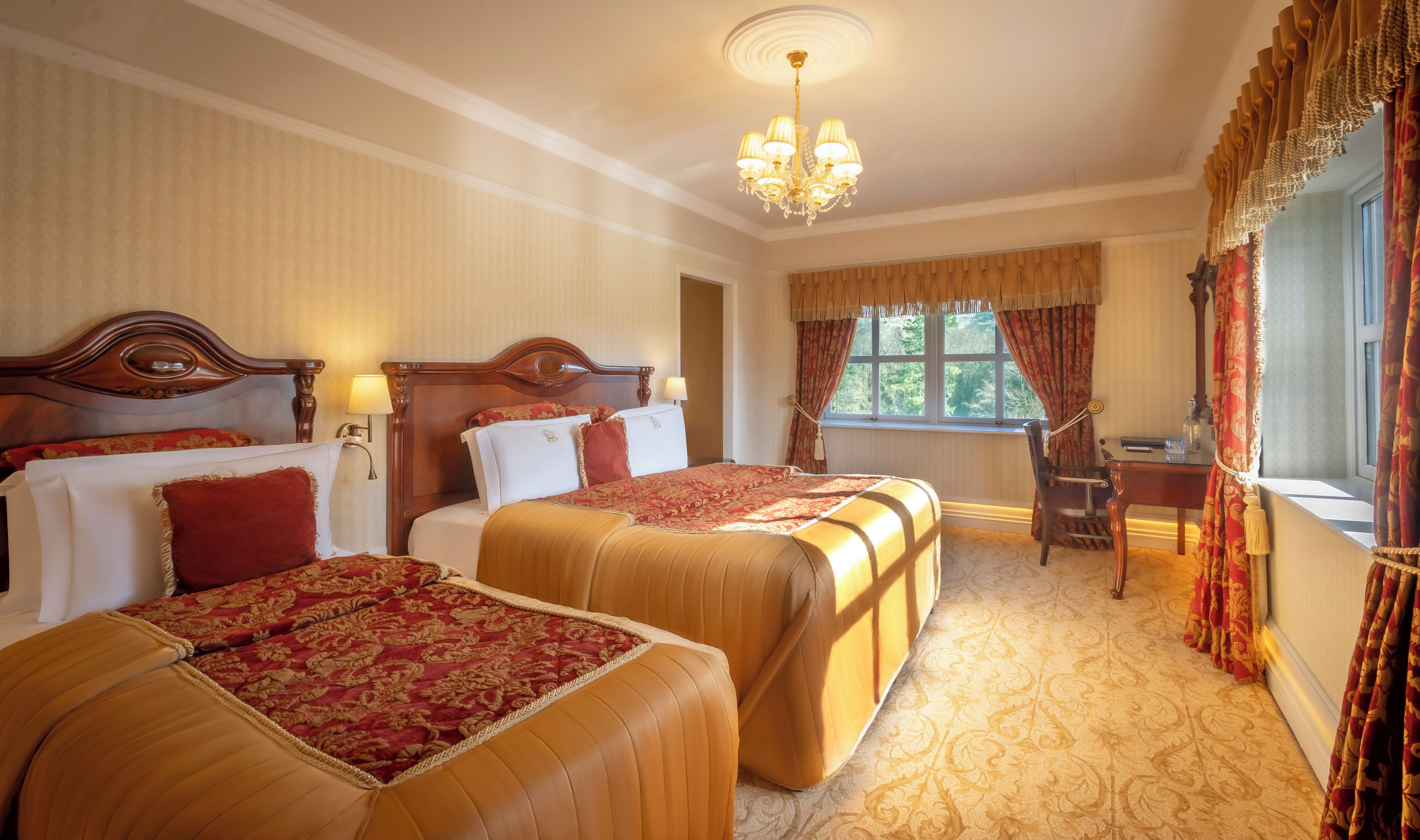 A hotel room with two beds, a chandelier, a desk, and a window with curtains.
