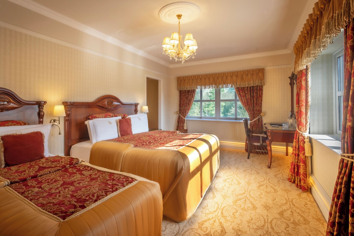 Habitación de hotel con dos camas, un candelabro, un escritorio y una ventana con cortinas.