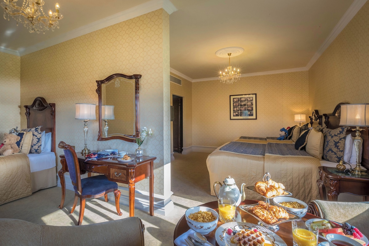 Habitación de hotel con dos camas, una mesa de comedor con desayuno, un rincón de estar con una silla y un candelabro.