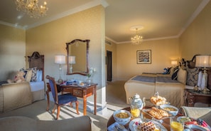 Habitación de hotel con dos camas, una mesa de comedor con desayuno, un rincón de estar con una silla y un candelabro.