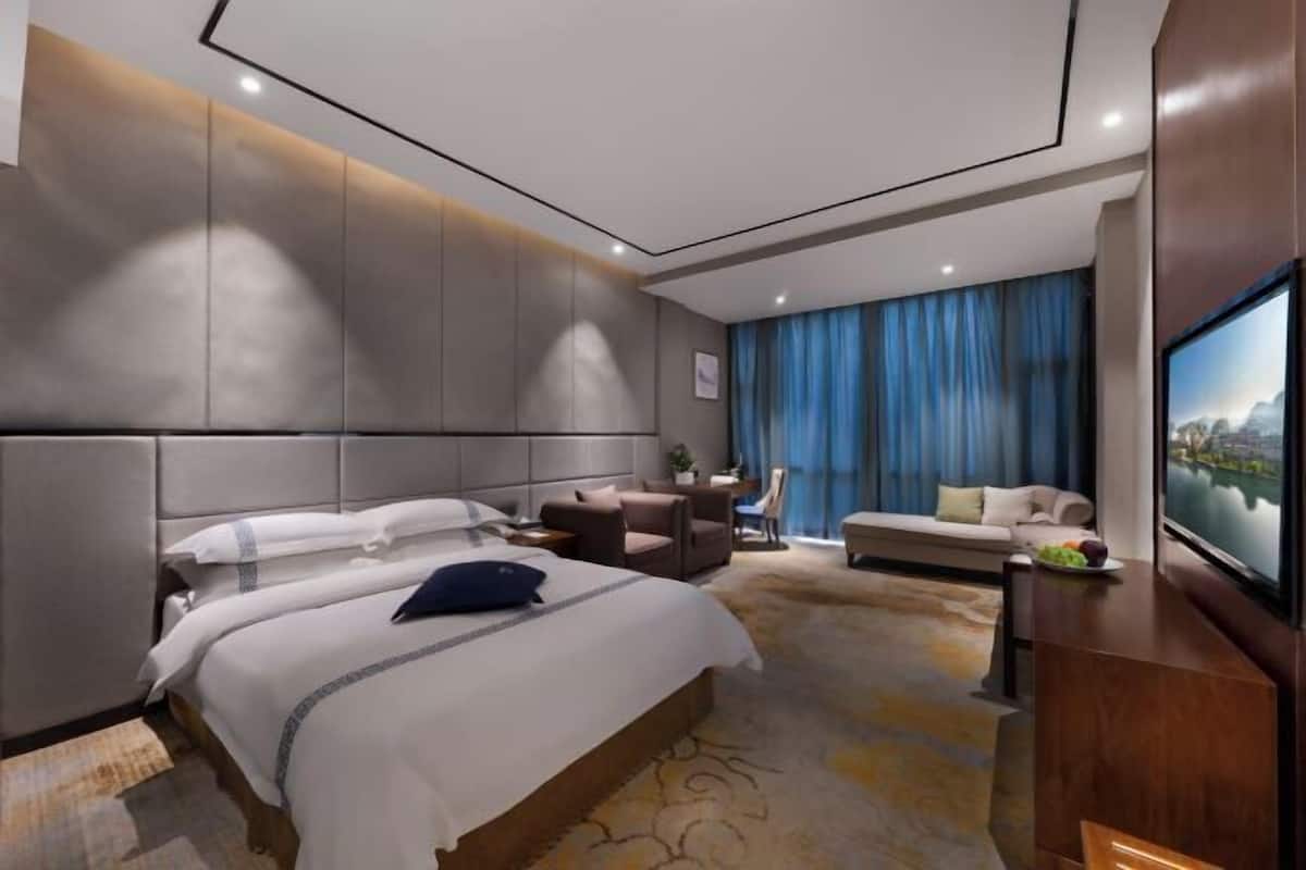 A modern hotel room with a large bed, a seating area with a sofa and chairs, a television, and a large window with curtains.