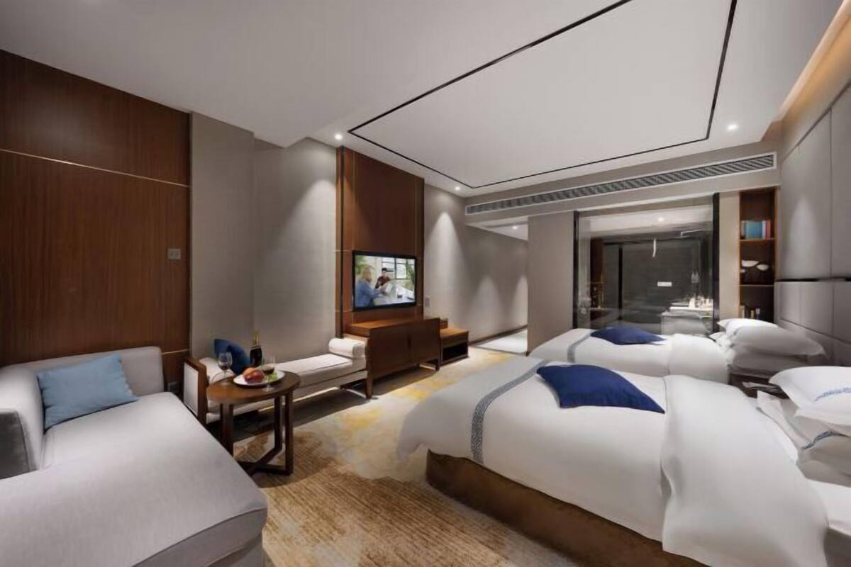 A modern hotel room with a sofa, two beds, a TV, a desk, and a bookshelf.