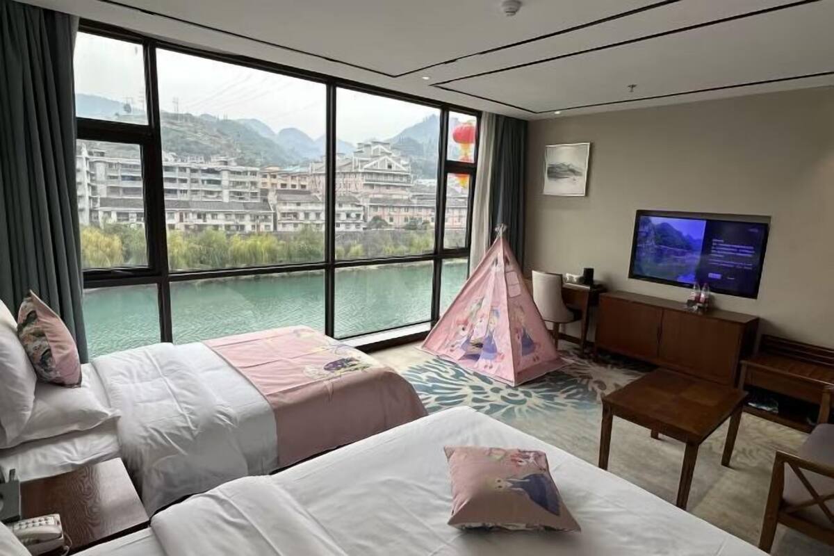 A hotel room with two beds, a TV, a desk, and a view of a building and mountains.