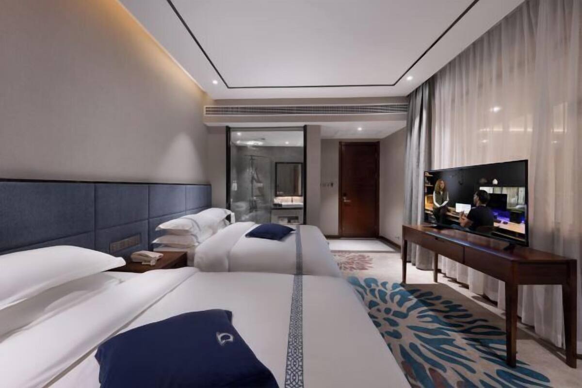 A hotel room with a bed, a TV, a desk, and a bathroom visible through a glass door.