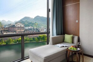 Una habitación de hotel moderna con un amplio ventanal que ofrece vistas a un río y edificios, un sofá con un libro y fruta, y una pequeña mesa con una copa de vino.