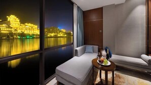 A modern hotel room with a large window, a sofa, a small table with a bottle and glasses, and a view of a lit-up building.