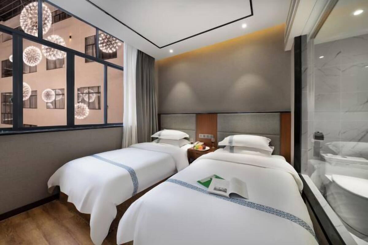 A hotel room with two beds, a large window with circular light fixtures, a bathroom with a marble wall, and a view of another building.