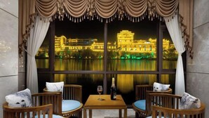 A room with four wooden chairs and a small table, a bottle of champagne, and a view of a building across a body of water.