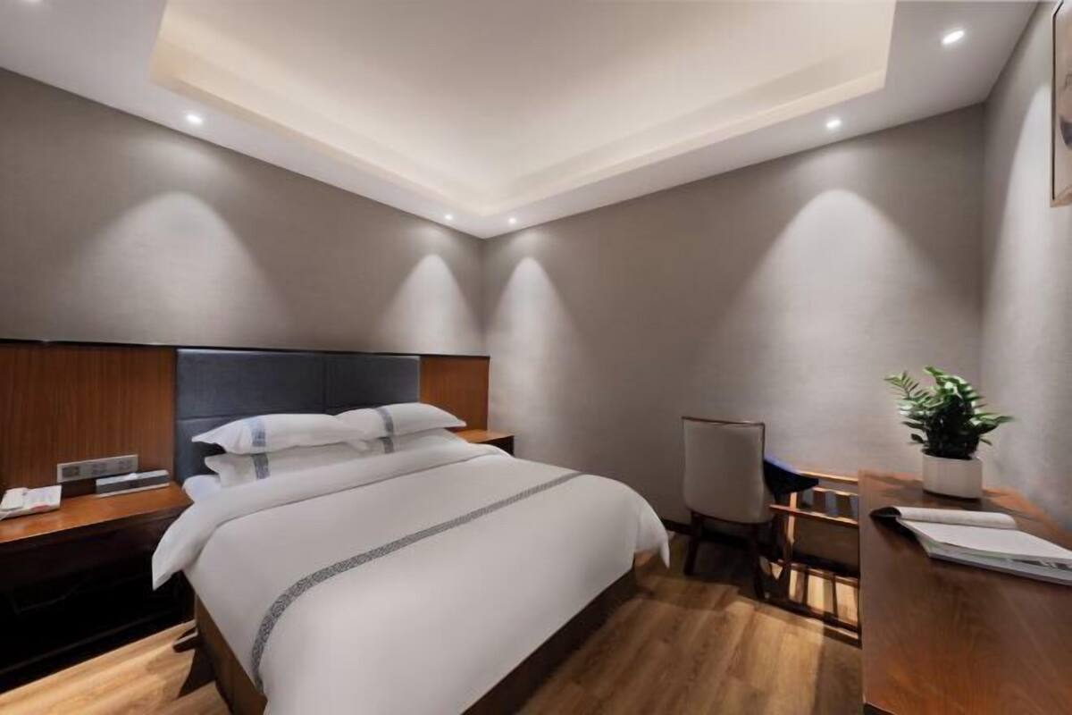 A modern hotel room with a bed, bedside table, desk, chair, and a small potted plant.