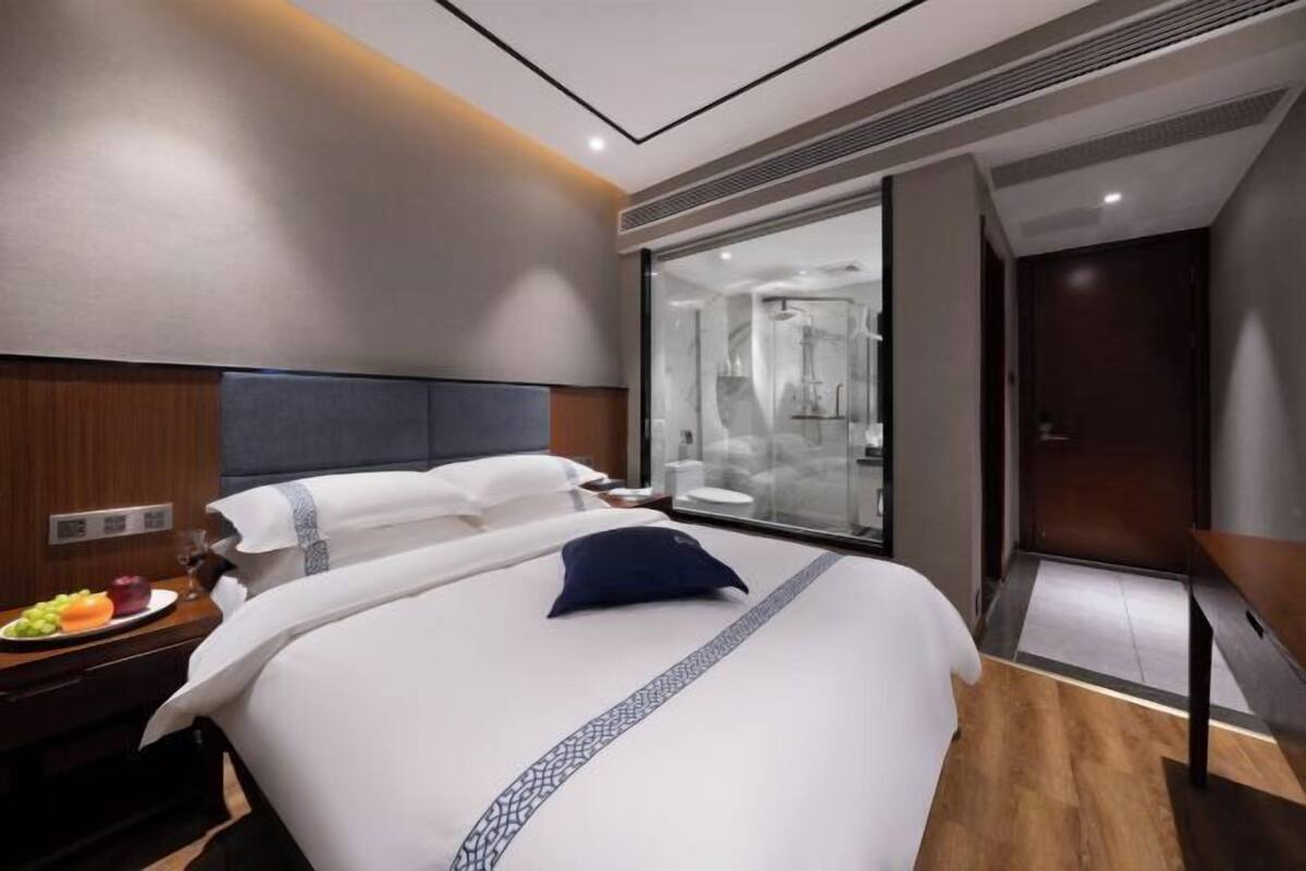A modern hotel room with a large bed, a bedside table with fruit, a glass shower enclosure, and a wooden floor.