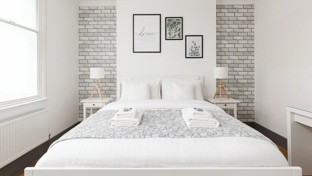 Quarto com parede de tijolos brancos, cama com roupa de cama branca, duas mesas de cabeceira com abajures e obras de arte emolduradas na parede.