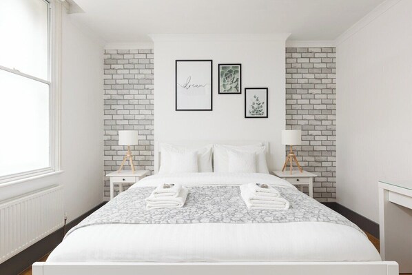 A bedroom with a white brick wall, a bed with white bedding, two bedside tables with lamps, and framed artwork on the wall. - New! Central 1BR, steps to Soho, Oxford St (Greater London)