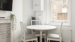 Uma sala de jantar clara com uma mesa redonda e quatro cadeiras, uma TV montada, um radiador branco e uma janela com vista para um edifício de tijolos.