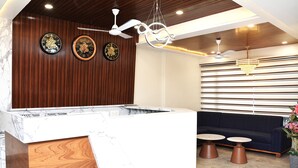 A reception area with a white counter, wooden panel wall, two clocks, ceiling fans, a chandelier, a dark blue sofa, and two round tables. - Hotel Aaradhya inn (Dwarka)