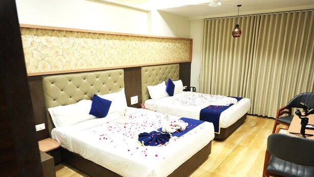 A hotel room with two beds, a wooden floor, a ceiling fan, and a window with curtains. - Hotel Aaradhya inn (Dwarka)