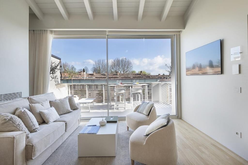 A modern living room with a sofa, armchair, and coffee table, a balcony with outdoor furniture, and a large window offering a view of buildings and trees.