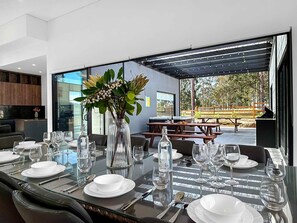 A modern dining area with a glass table, white plates, and clear glasses. There is a large window with a view of the outdoors. - Sereno - Fabulous golf resort getaway (POKOLBIN)