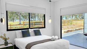 Een moderne slaapkamer met een groot bed, uitzicht op een omheind buitengebied en een klein nachtkastje met een vaas.