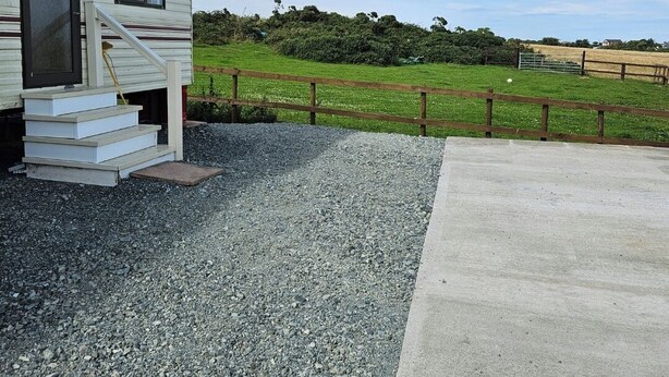 Uma casa móvel com um pátio de concreto, entrada com pedrisco e um campo verde ao fundo.