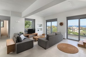 Ein modernes Wohnzimmer mit grauen Sofas, einem Flachbildfernseher, einem Holztisch und großen Fenstern mit Meerblick.