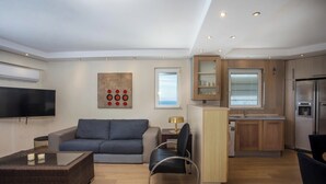 Ein modernes Wohnzimmer mit einer Couch, einem Couchtisch und einem Fernseher. Es gibt eine Küchenzeile mit Holzschränken und einem Kühlschrank.