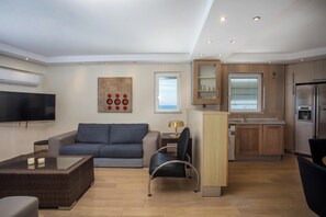 A modern living room with a sofa, a coffee table, and a TV. There is a kitchen area with wooden cabinets and a refrigerator. - Molos Seaside Apartment in Rhodes Town (Rhodes)