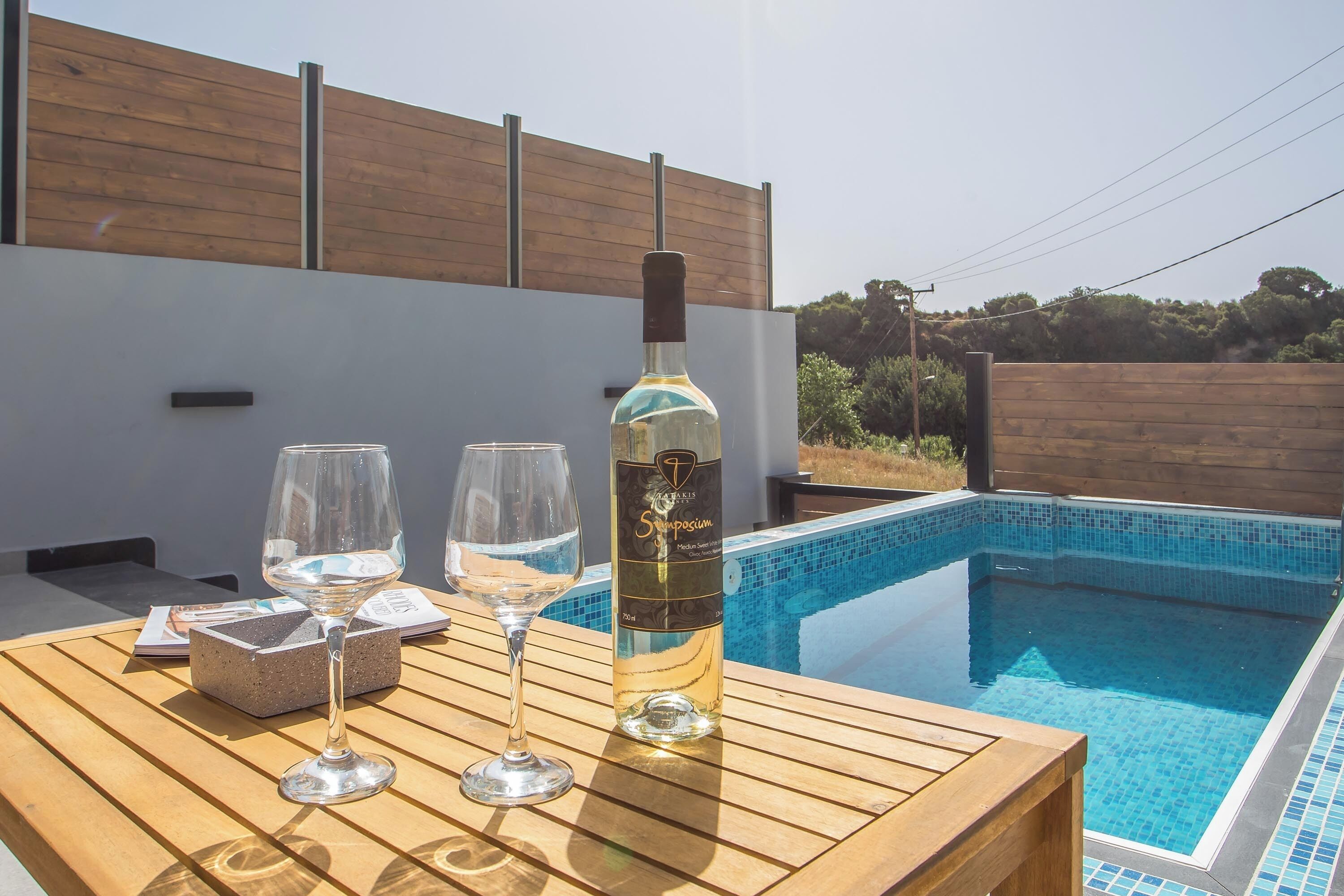 Une table en bois, avec deux verres à vin et une bouteille de vin, près d’une piscine.