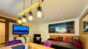 Ein modernes Wohnzimmer mit einem Holzesstisch, einer Couch mit Kissen, einem Fernseher und einem Gemälde von einer Berglandschaft.