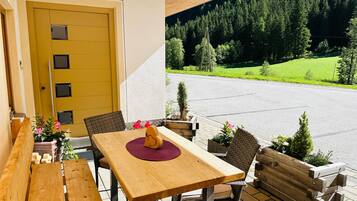 An outdoor seating area with a wooden table and chairs, a yellow door, and a view of a road and forest.