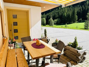 Un área de estar al aire libre con una mesa y sillas de madera, una puerta amarilla y vistas a una carretera y un bosque.