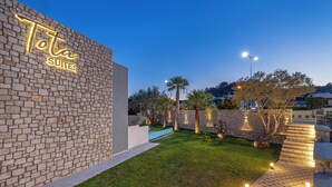 A hotel exterior with a brick wall, a neon sign reading "Tota Suites," a well-maintained lawn, and stone steps leading to the entrance. - Tota Suites - Suite 3 (Rhodes)