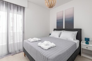 Ein ordentlich bezogenes Bett mit weißen Leinen, einem grauen Kopfteil und einem Nachttisch mit einer blauen Lampe.