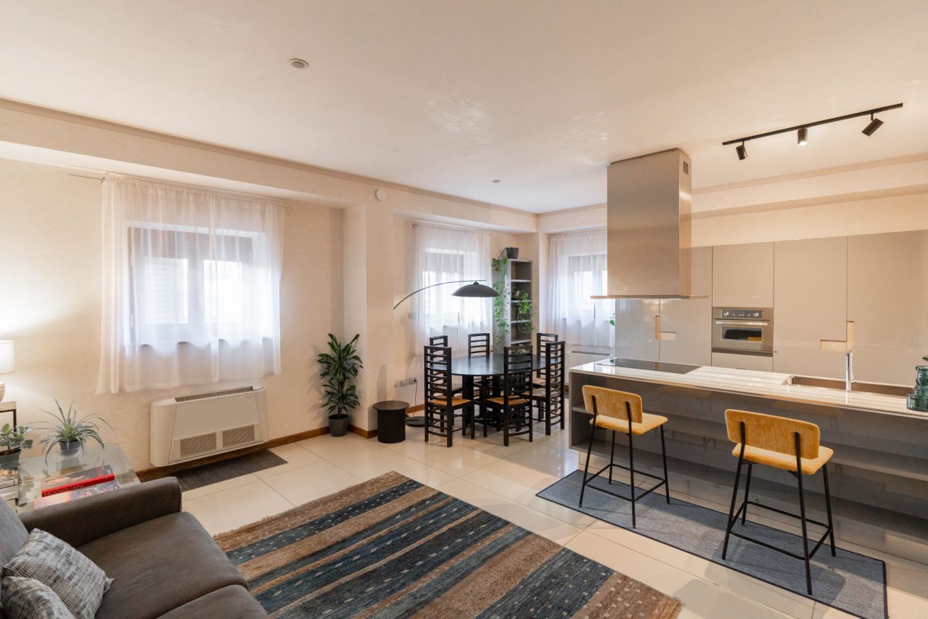 Una sala moderna con un sofá, una mesa de comedor y sillas, una zona de cocina con estufa y armarios, y un ventanal con cortinas translúcidas.