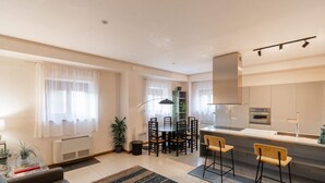 Ein modernes Wohnzimmer mit einer Couch, einem Esstisch und Stühlen, einer Küchenzeile mit Herd und Schränken sowie einem großen Fenster mit durchsichtigen Vorhängen.