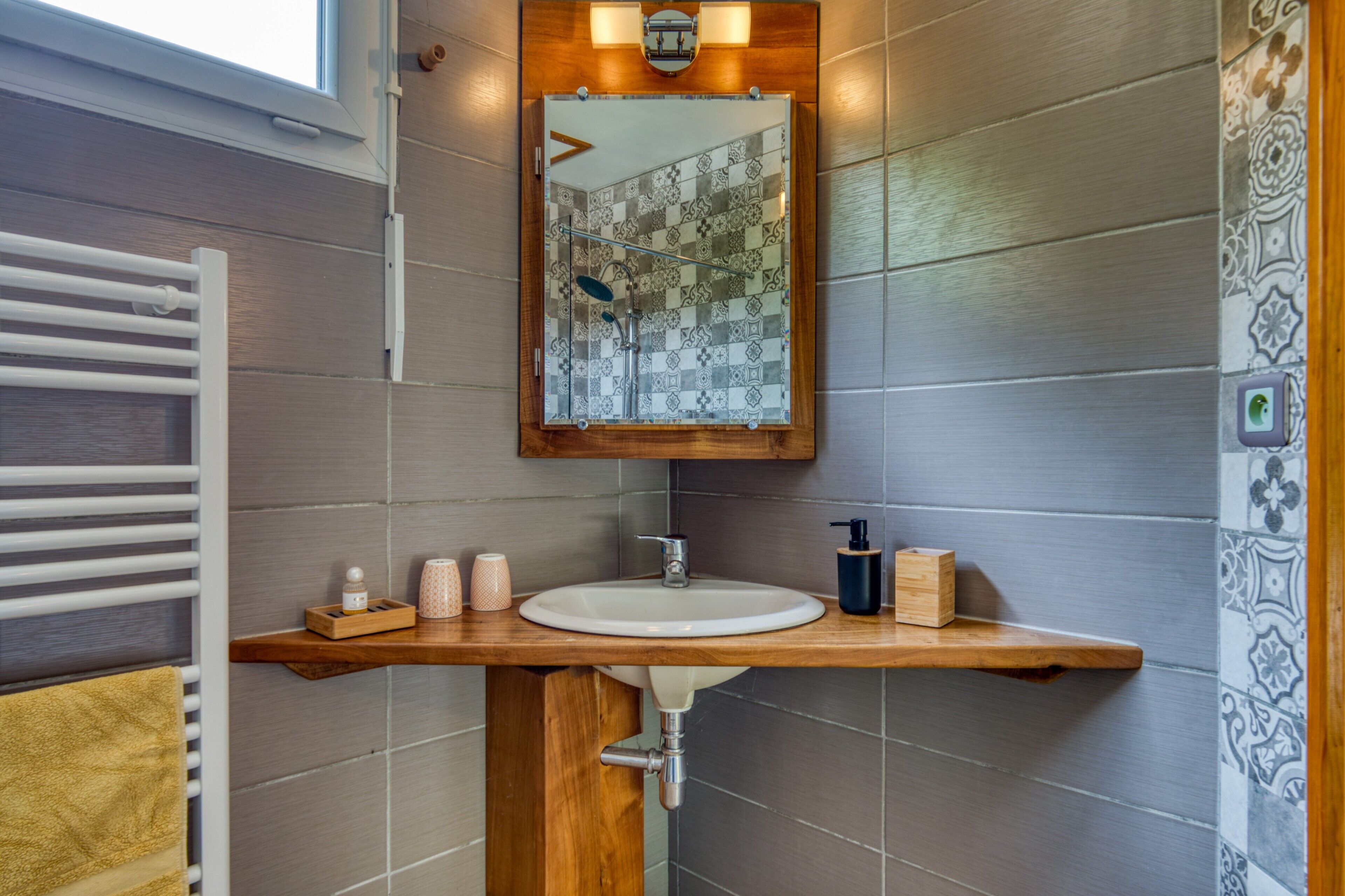 Un baño con tocador de madera, un lavabo blanco, un espejo con marco de madera y azulejos estampados.