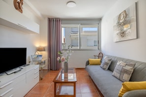 A modern living room with a flat-screen TV, a glass coffee table, a sofa with cushions, a vase with flowers, and a painting on the wall.