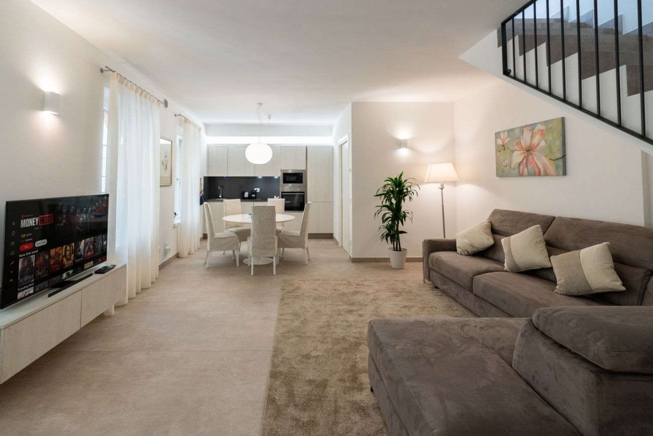 A modern living room with a flat-screen TV, a sofa, a dining area, and a staircase.