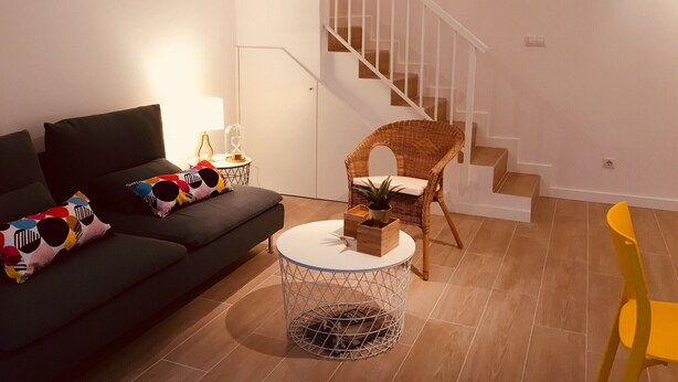 Ein Wohnzimmer mit einer Couch, einem runden Couchtisch, einem Korbstuhl, einem gelben Sessel und einer Treppe.