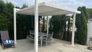 Ein überdachter Patio-Bereich mit Tisch und Stühlen, einem Liegestuhl und einer weißen Pergola.