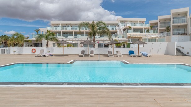 A swimming pool with lounge chairs and umbrellas, surrounded by a building with multiple balconies and a clear sky. - Vacation Apartment 'Casa Amor Costa Teguise' with Wi-Fi (Costa Teguise)