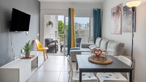 A modern living room with a sofa, dining table, and chairs, a flat-screen TV mounted on the wall, and a balcony with a view. - Vacation Apartment 'Casa Amor Costa Teguise' with Wi-Fi (Costa Teguise)